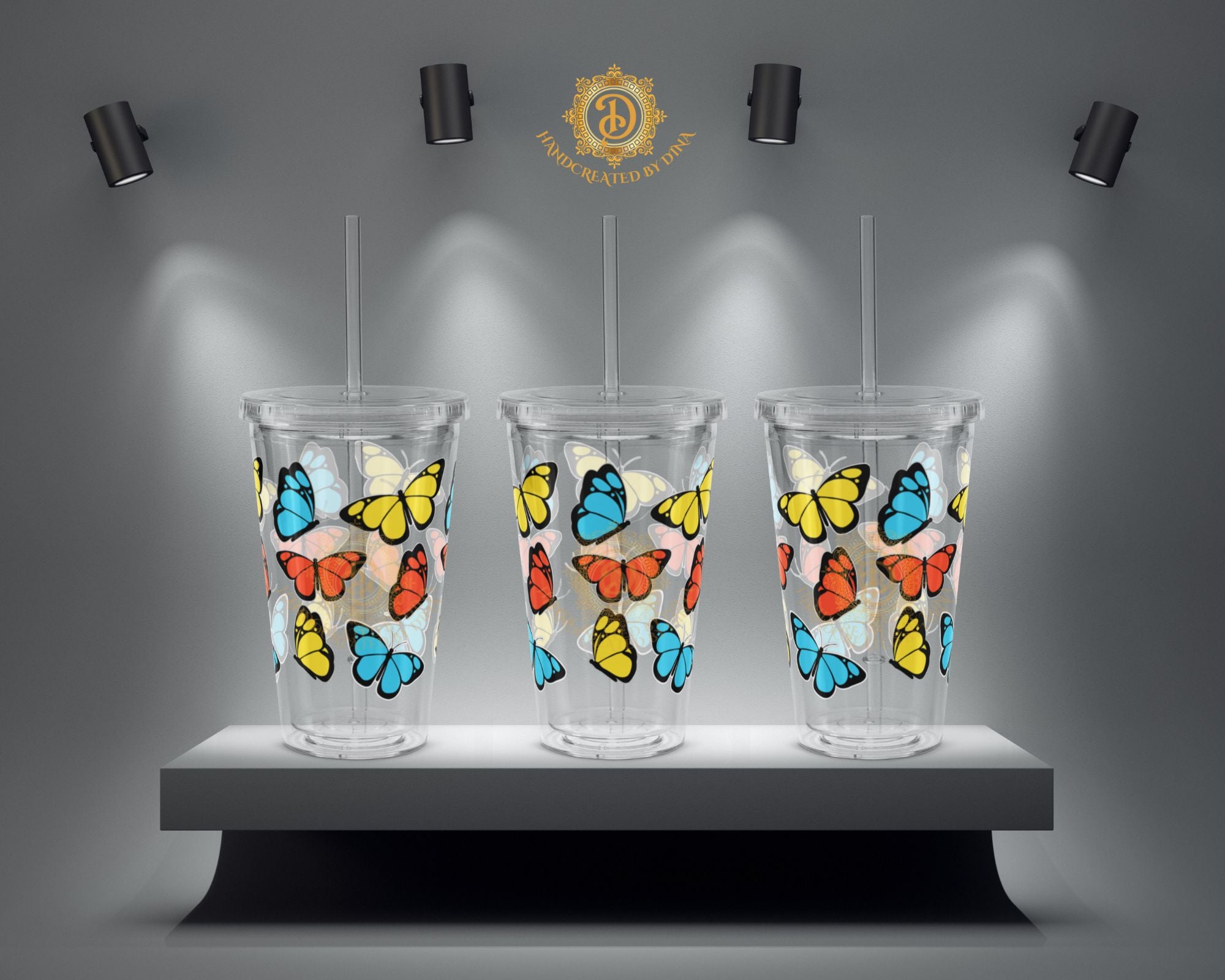 Three clear tumblers with colourful butterfly designs on a grey platform with spotlights.