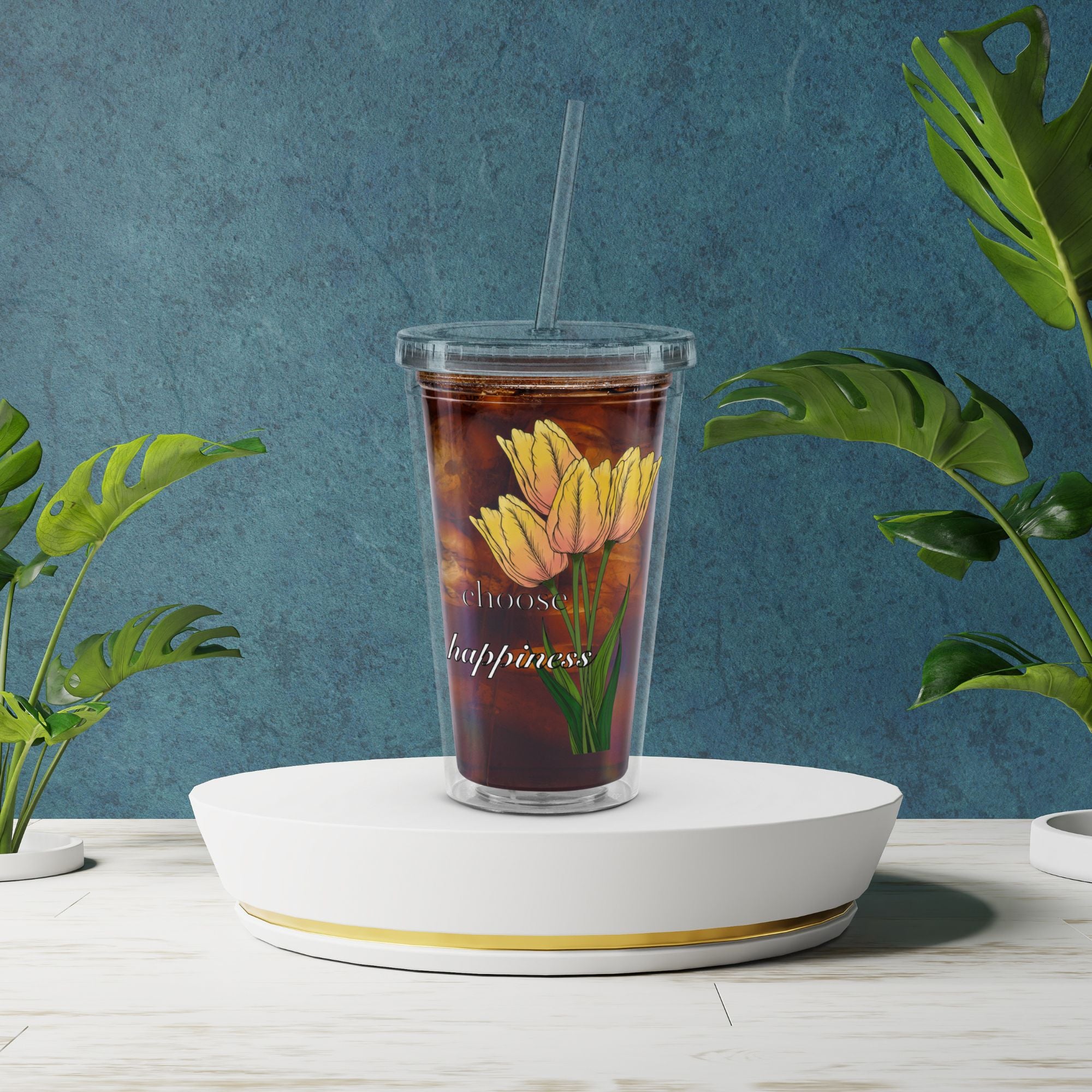 Tumbler with floral design and text on a white stand against a blue wall with plants.