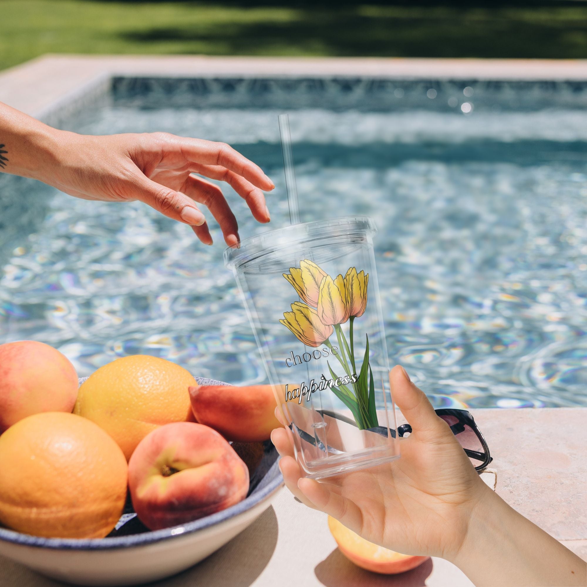Person holding a tumbler with tulip design by a poolside with fruits on a table.
