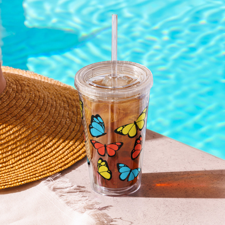 A clear plastic tumbler with colourful butterfly designs, accompanied by a straw, placed on a surface near a poolside. A straw hat is also visible in the background.