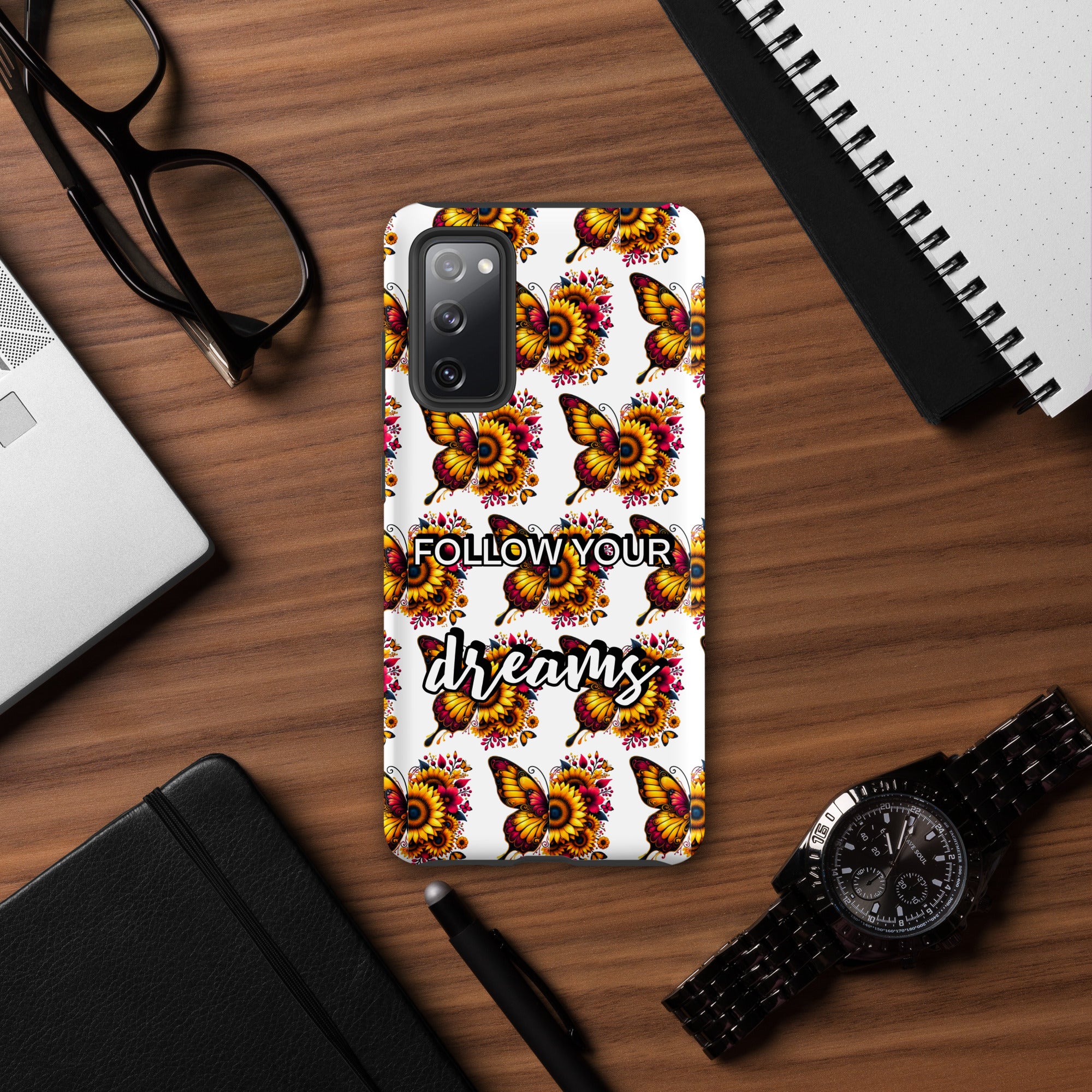 Phone case with floral design and 'Follow Your Dreams' text on a wooden desk with accessories.