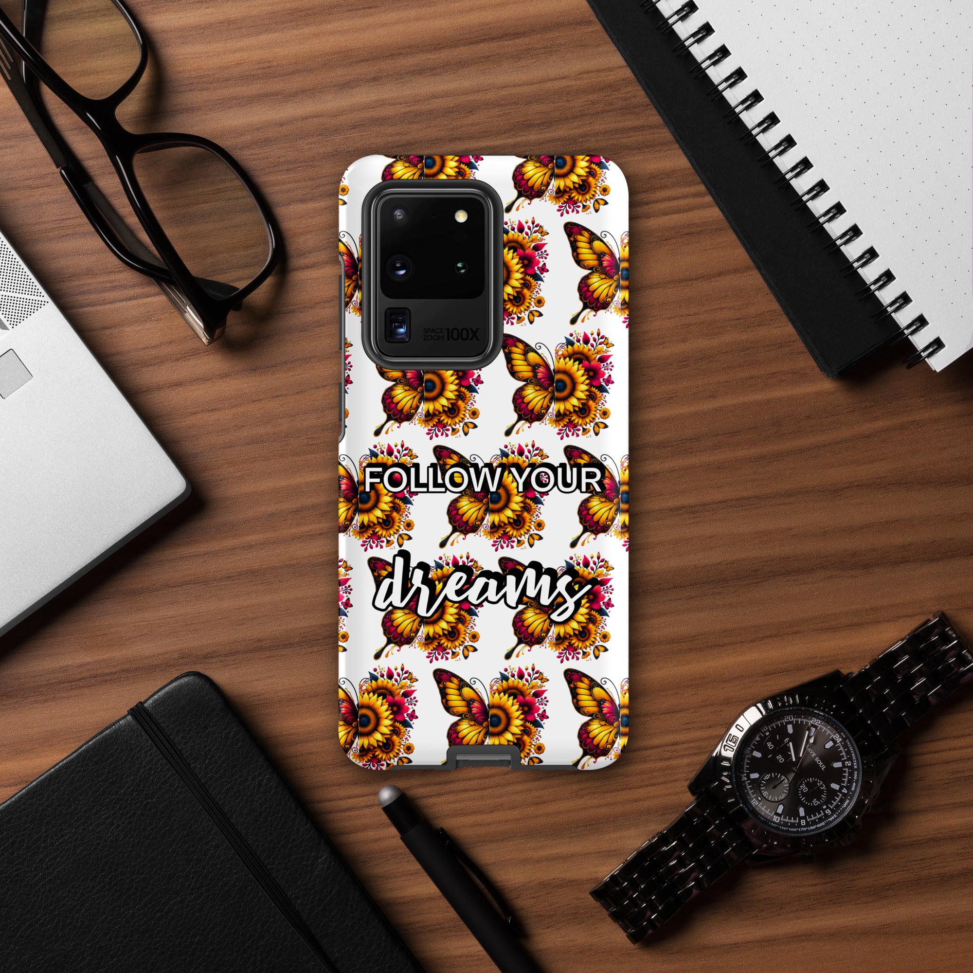 Phone case with butterfly design and 'Follow Your Dreams' text on a wooden desk.