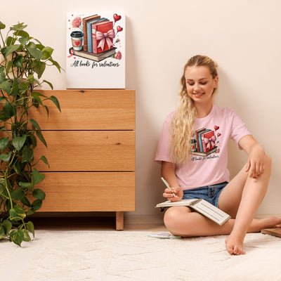 Woman sitting on the floor with a book, wearing a pink t-shirt with a design, in a room with a wooden dresser and plant.