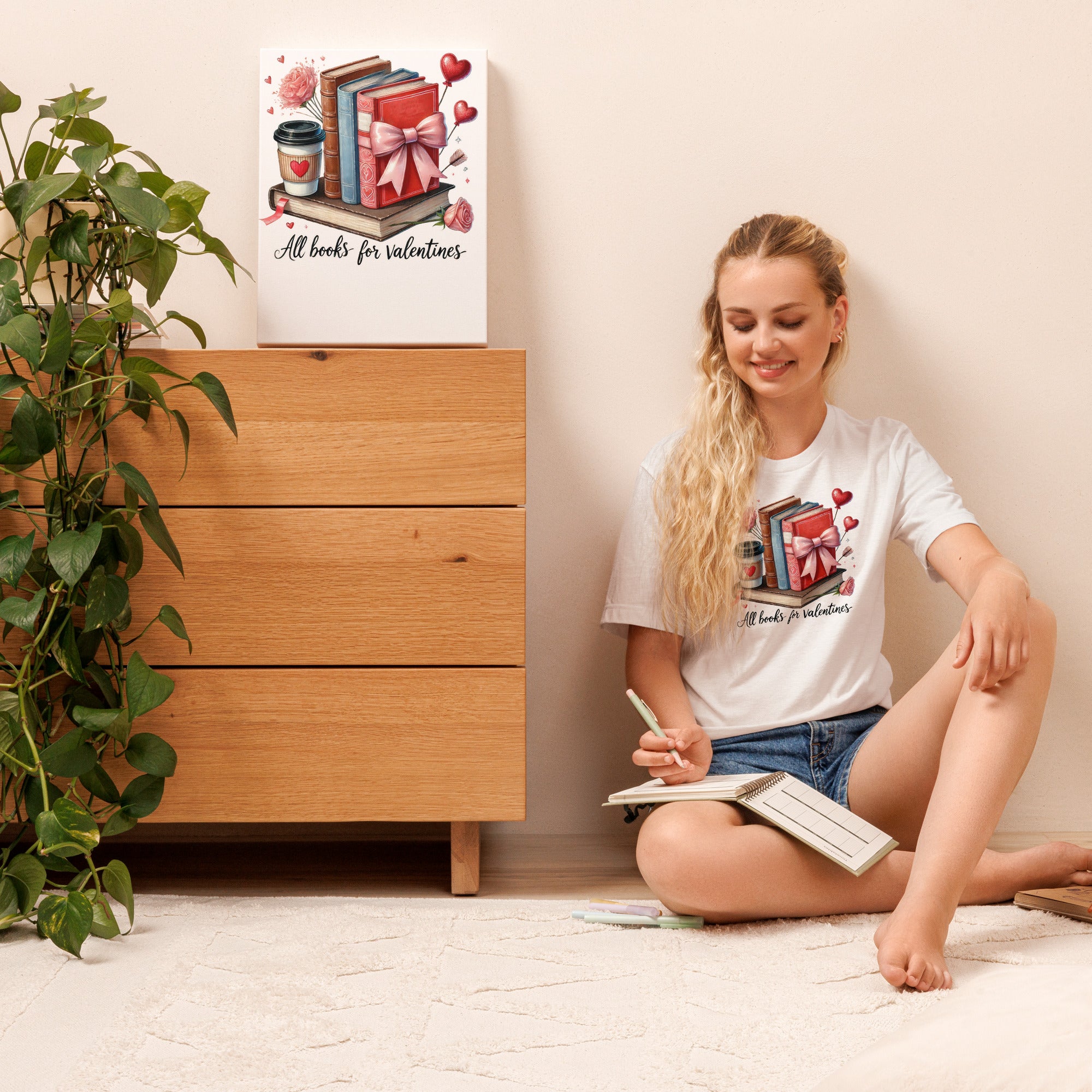Woman sitting on the floor with a book, wearing a white t-shirt with a design, in a room with a wooden dresser and plant.