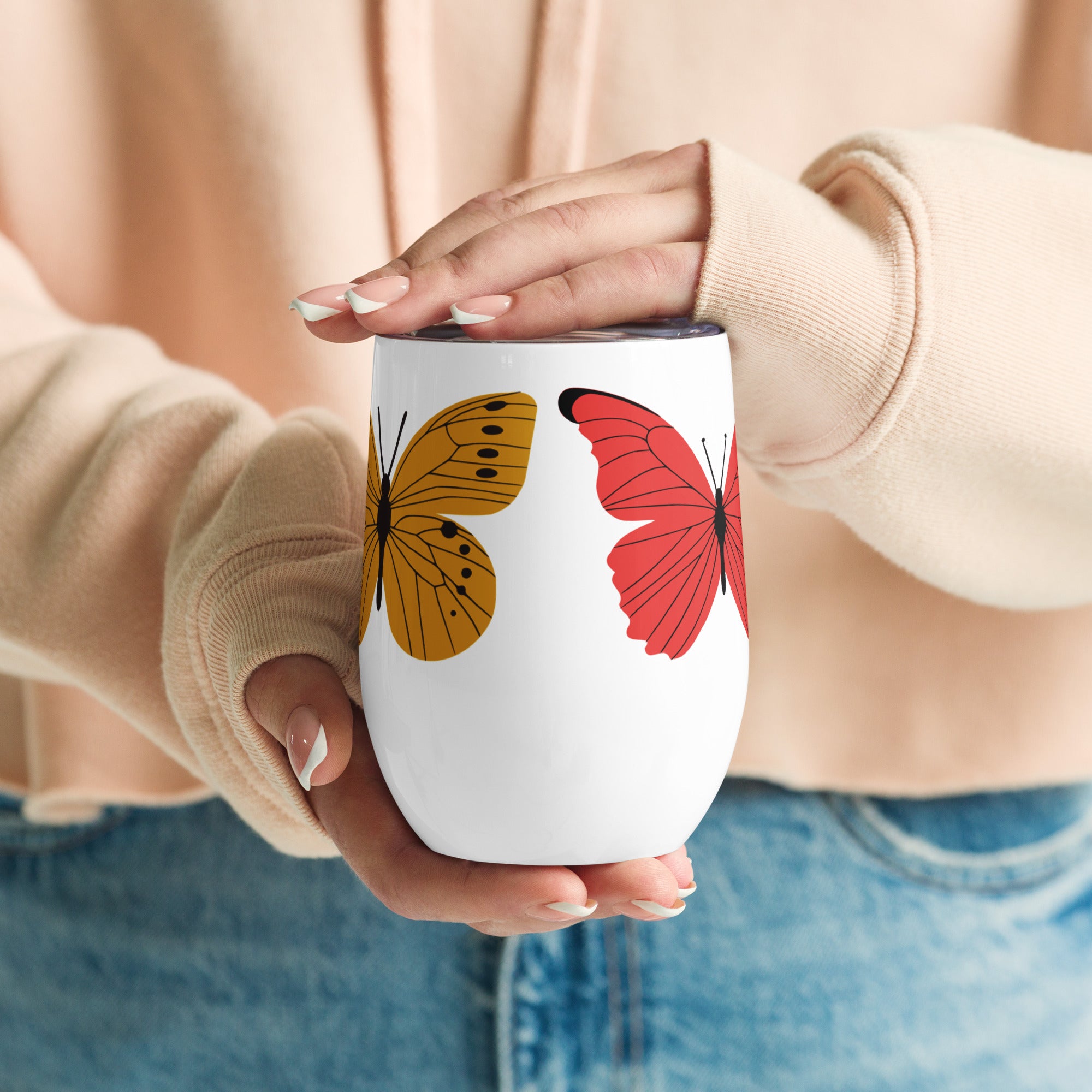 Person holding a white wine mug with butterfly designs, wearing a beige sweater and blue jeans.