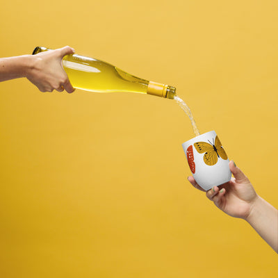 Person pouring white wine from a bottle into a white cup with a butterfly design on a yellow background