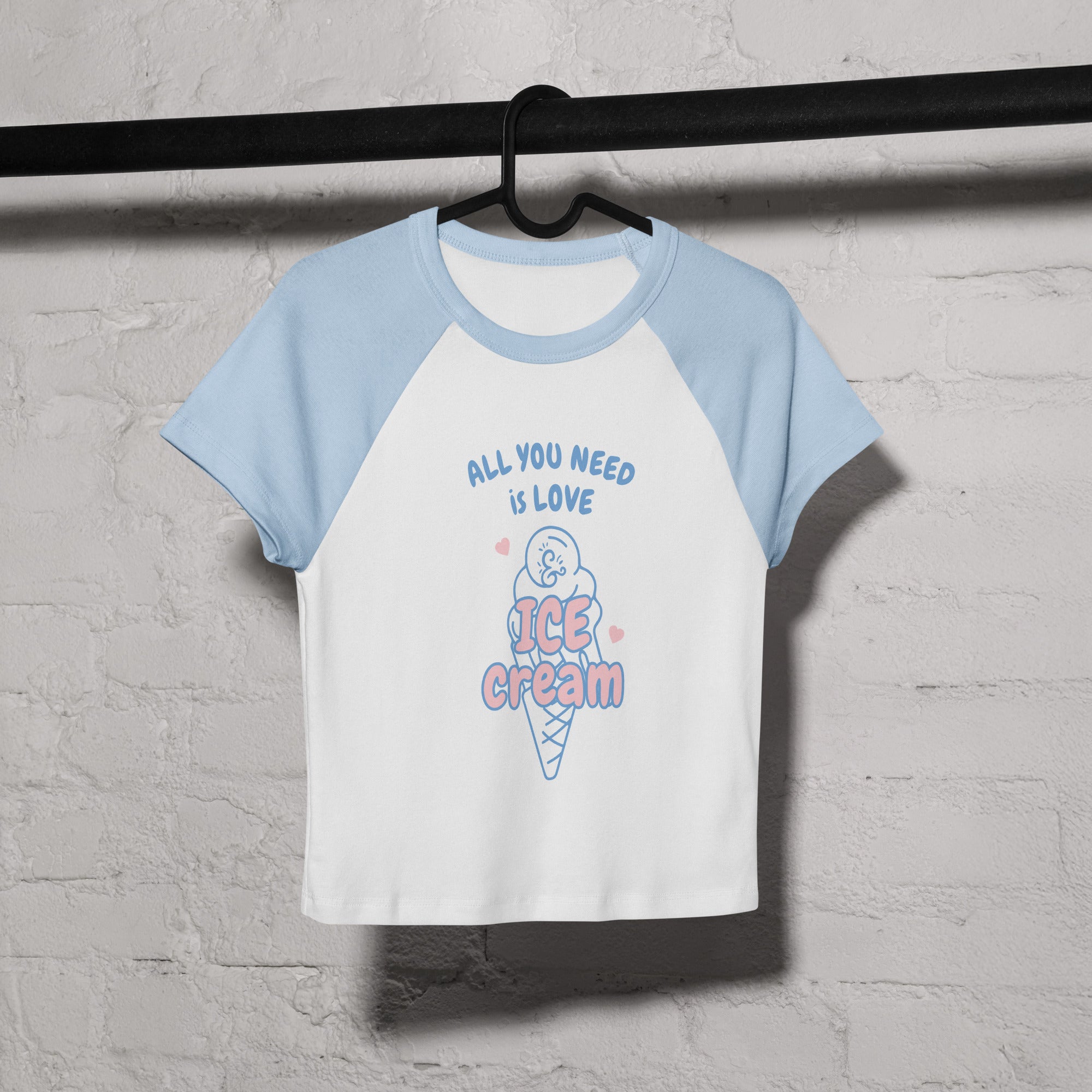 White and light blue t-shirt with ice cream graphic and text on a hanger against a gray brick wall.