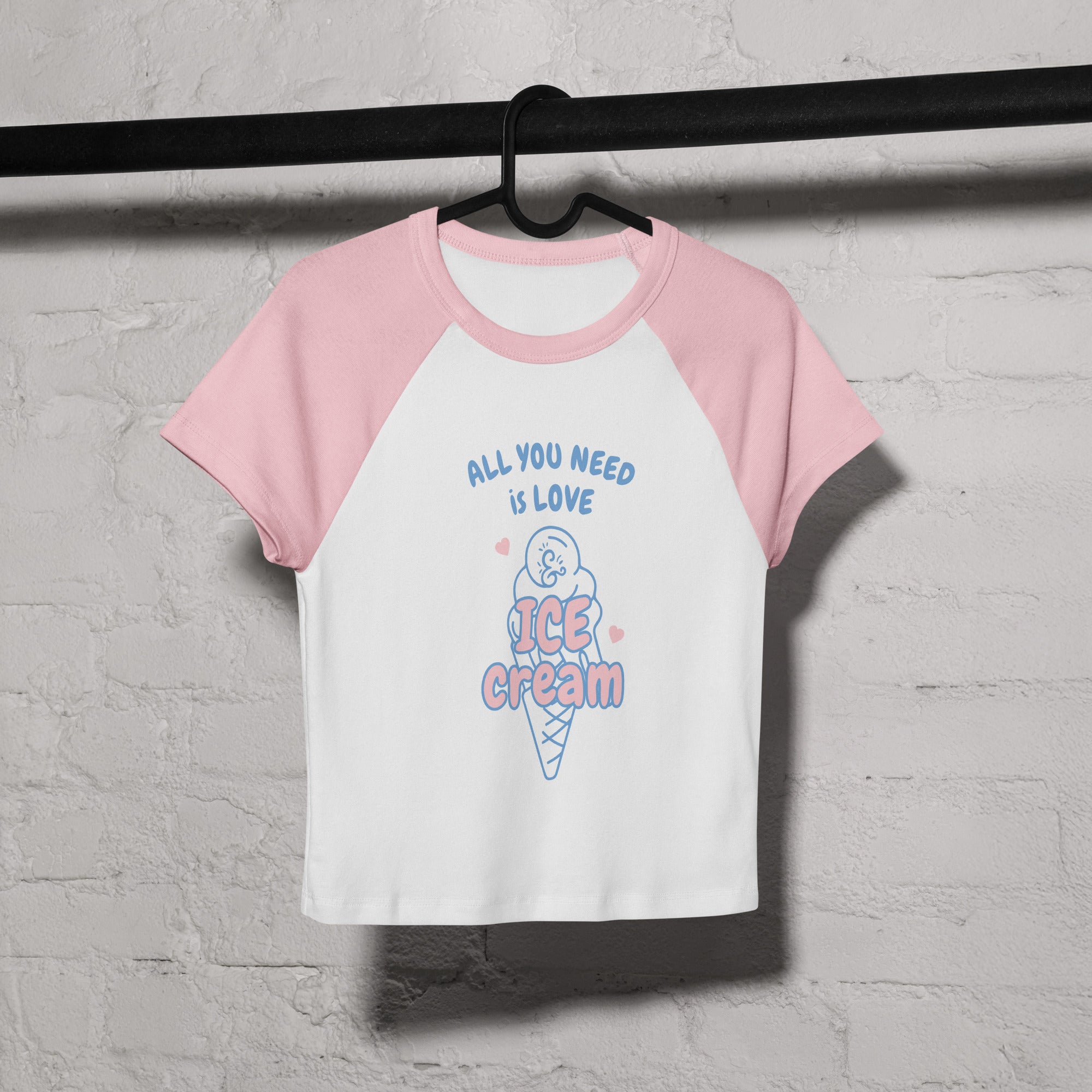 Pink and white baseball shirt with ice cream graphic and text on a hanger against a gray brick wall.