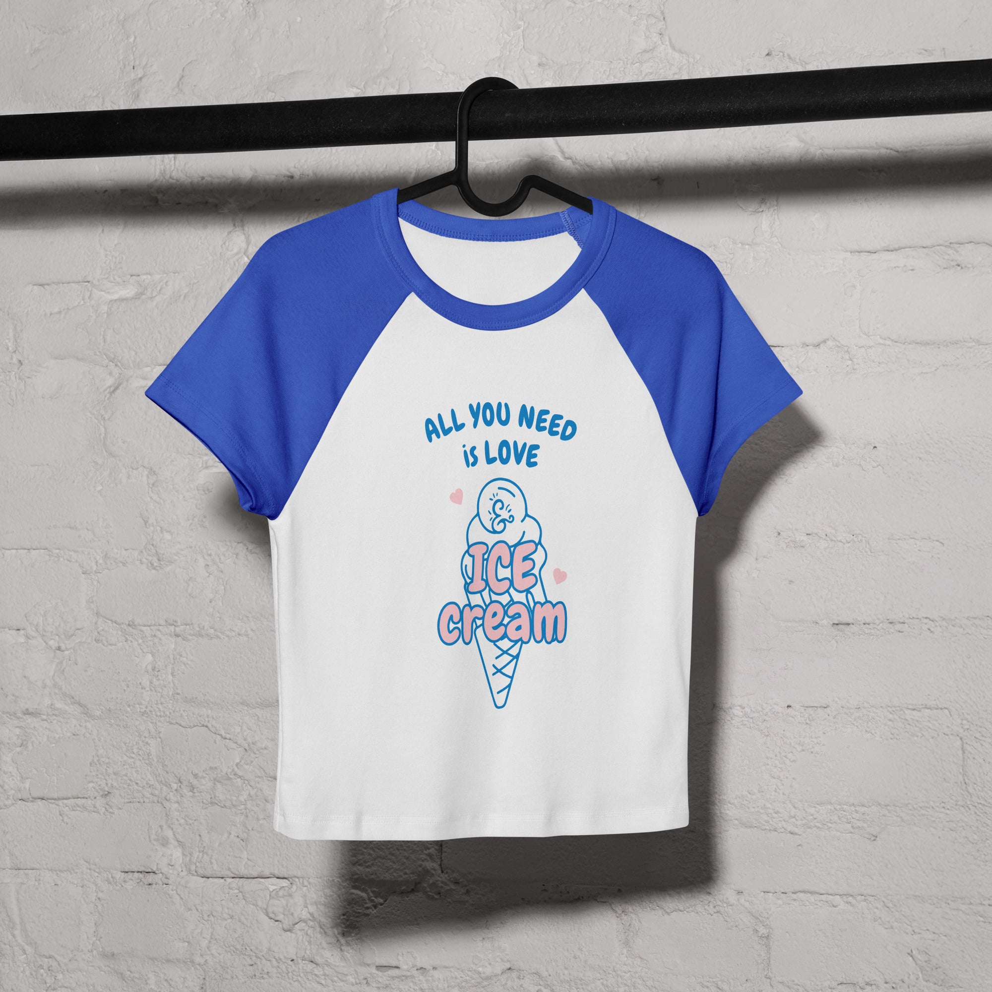 White and blue t-shirt with 'All you need is love and ice cream' text on a hanger against a gray brick wall.